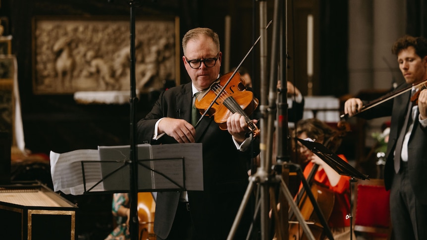 A man wearing a black suit and glasses plays the viola.