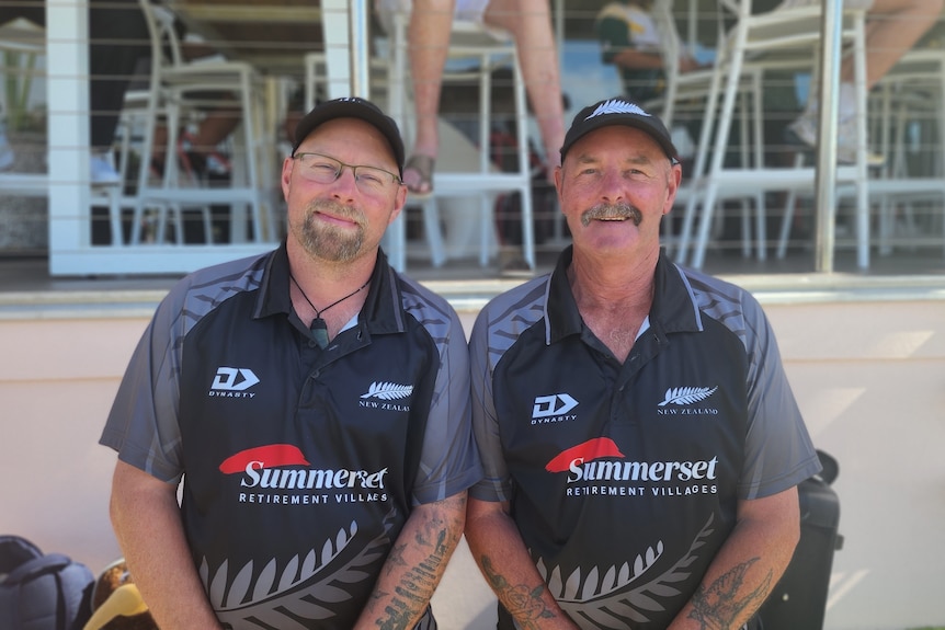 Two men sit on an outdoor bench in New Zealand sports shirts, smiling at the camera.