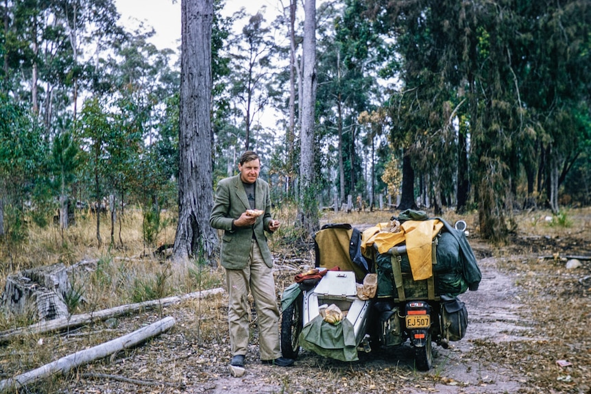 A man in a suit stands in the Australian bush next to a motorcycle with a sidecar.
