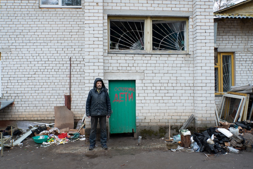 A man in a hooded puffer jacket stands outside a wrecked building looking stony-faced 