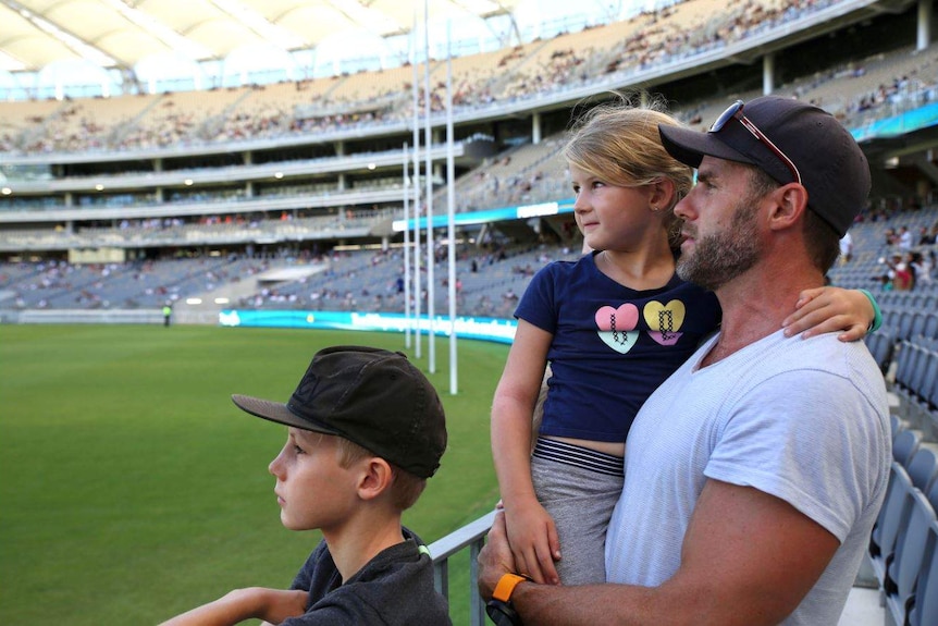 Mark holds Eloise on his hip, and Noah stands in front at the edge of the stadium ground.