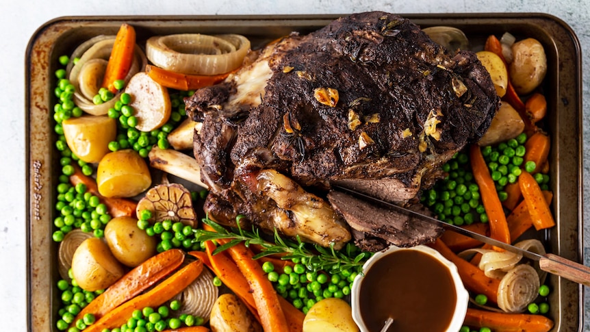 A lamb leg on a tray surrounded by baby carrots, potatoes, onion, peas and gravy, made easily in a slow cooker.