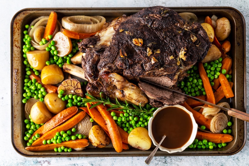 A lamb leg on a tray surrounded by baby carrots, potatoes, onion, peas and gravy, made easily in a slow cooker.