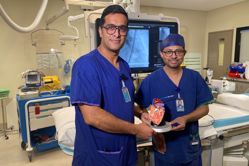 Two doctors stand in the middle of a hospital room holding a model of a heart.