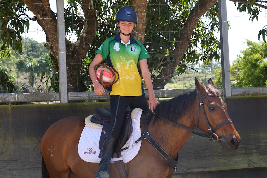 Katie Maund stands upright off the saddle on a horse, she holds a ball in a leather harness with six handles