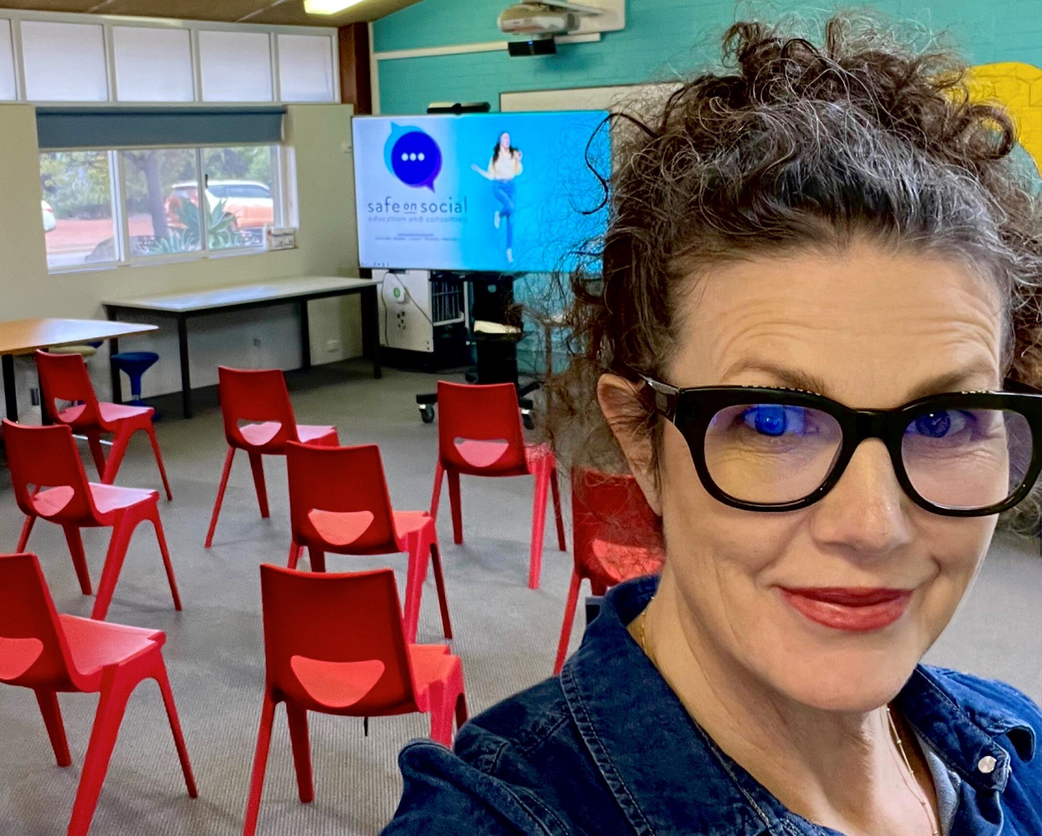 A middle-aged woman smiles as she takes a selfie in an empty classroom.