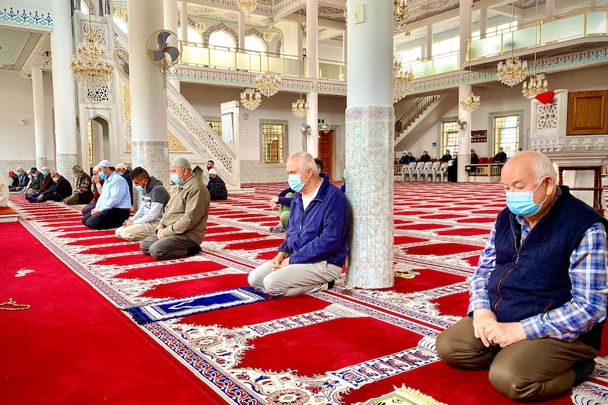 A group of men wearing face masks kneel on a carpet inside a mosque. 