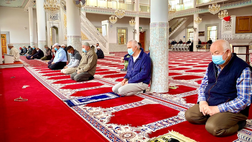 A group of men wearing face masks kneel on a carpet inside a mosque. 