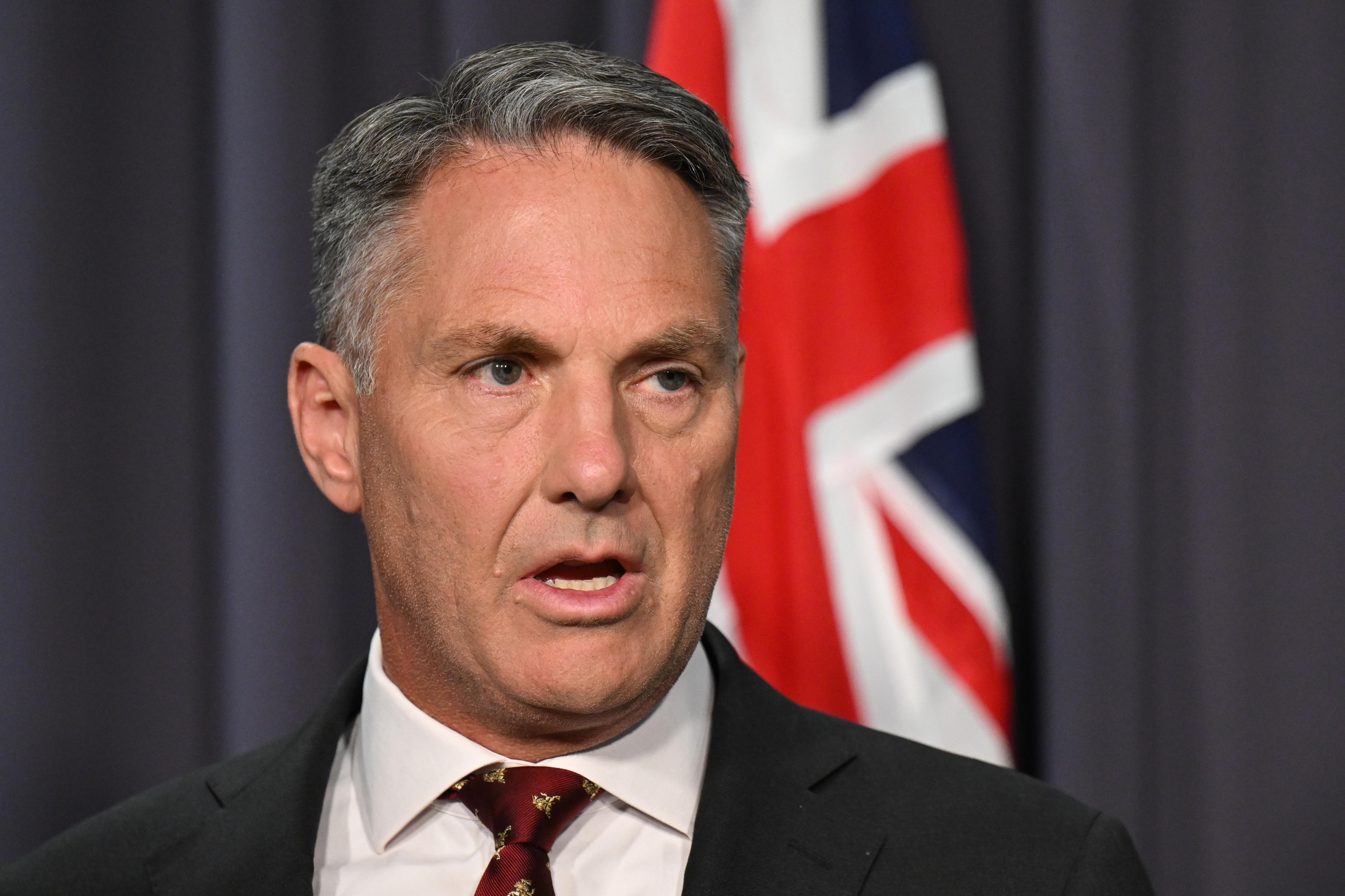 A close up of a white middle aged man in a suit and tie standing in front of a dark blue backdrop and Australian flag.
