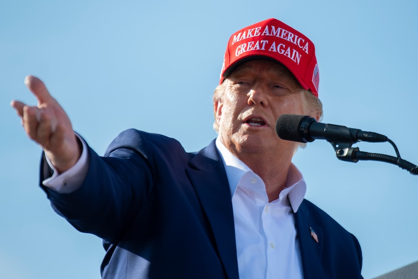 Donald Trump points to his right while standing in front of a microphone and wearing a Make America Great Again red cap