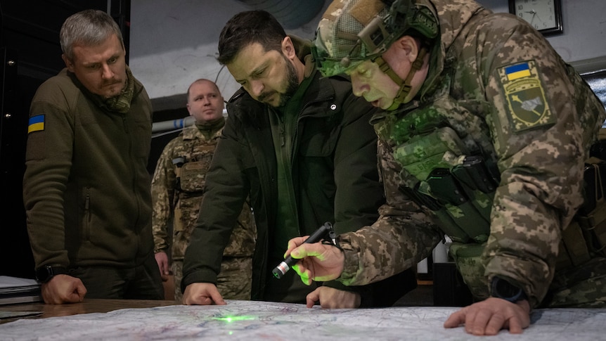 A group of four men, two in military uniforms and one wearing a helmet, lean over a table while studying a map.
