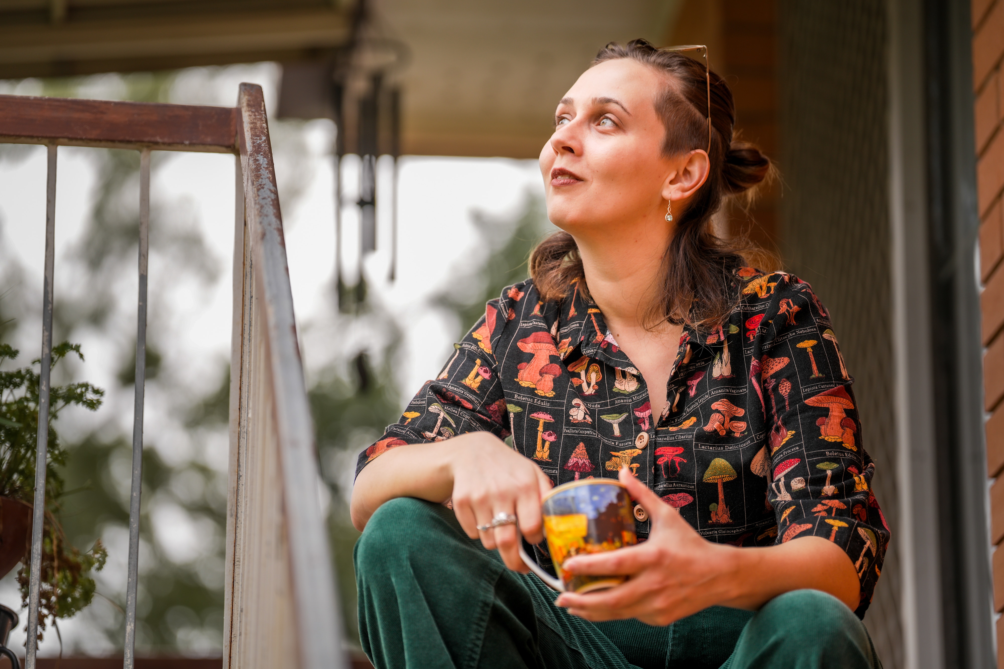 A woman with a colourful shirt holding a cup of tea while sitting on a staircase outside