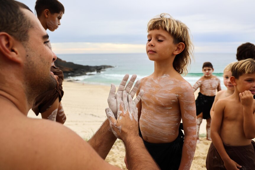 A young Aboriginal boy stands with his eyes closed preparing to be painted traditionally