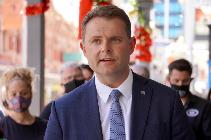 A man in a suit, white shirt and blue tie answers a question at a press conference