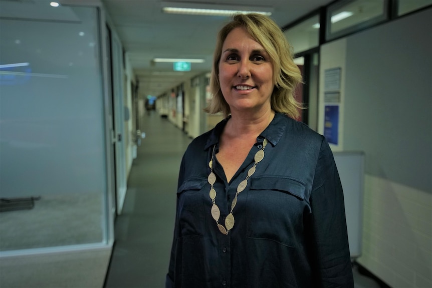A smiling woman with short blonde hair wearing a navy blue shirt and a long necklace stands in a corridor