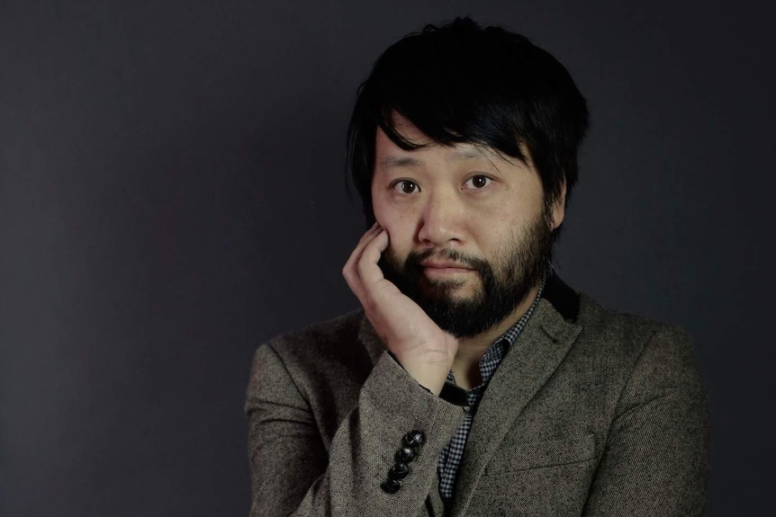 A man rests his head on a hand in front of a dark grey wall