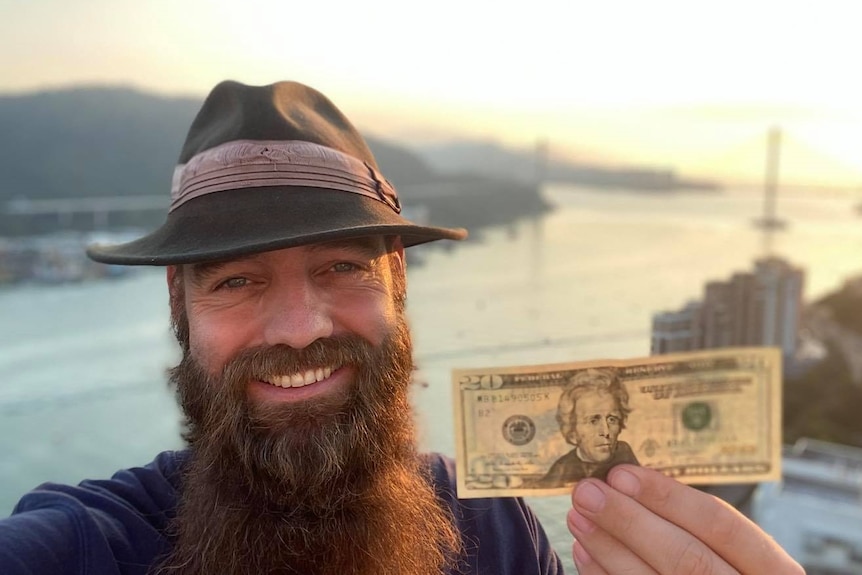 Bearded Thor, porte un chapeau, sourit en tenant un billet de 20 dollars américains avec une rivière ensoleillée derrière lui. 