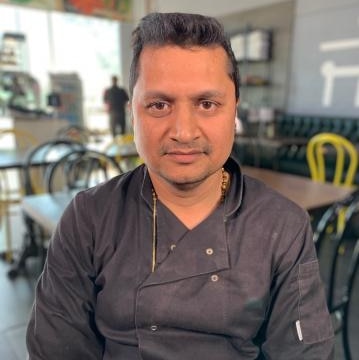 Tushar Patel sits in a restaurant he manages with dining table and chairs in the background.