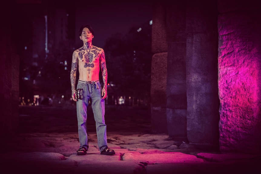 A young Korean man stands shirtless, exposing his heavily tattooed chest in a laneway at night 