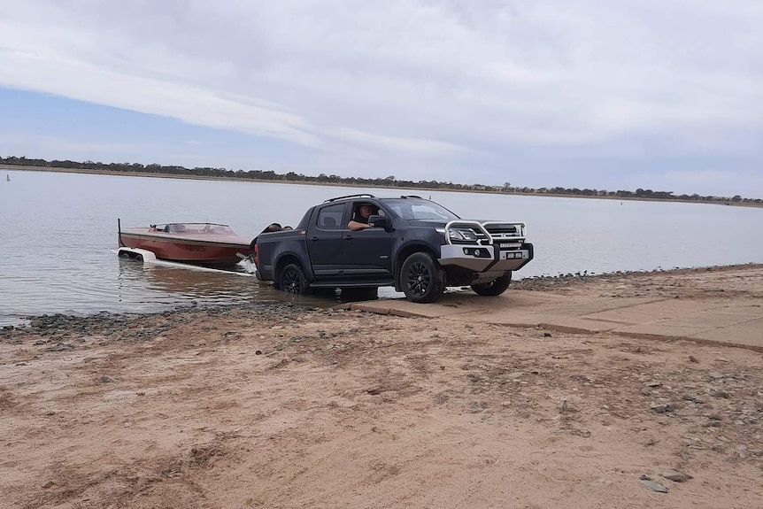 A ute pulls a boat on a trailer out of a lake.