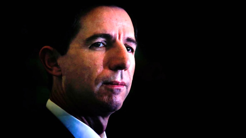 Simon Birmingham stands in the shadows and looks to his right to the media.