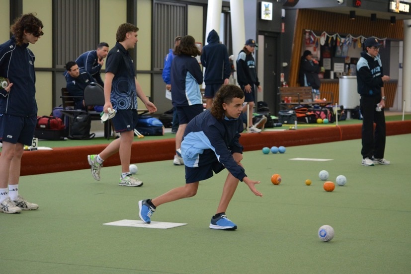 A boy with a mullet rolling a ball at a lawn bowls tournament.