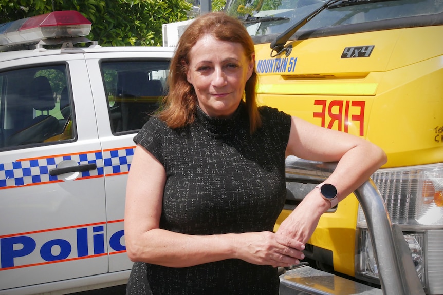 A middle-aged, ginger-haired woman in a dark dress leans on a fire truck near a police vehicle.