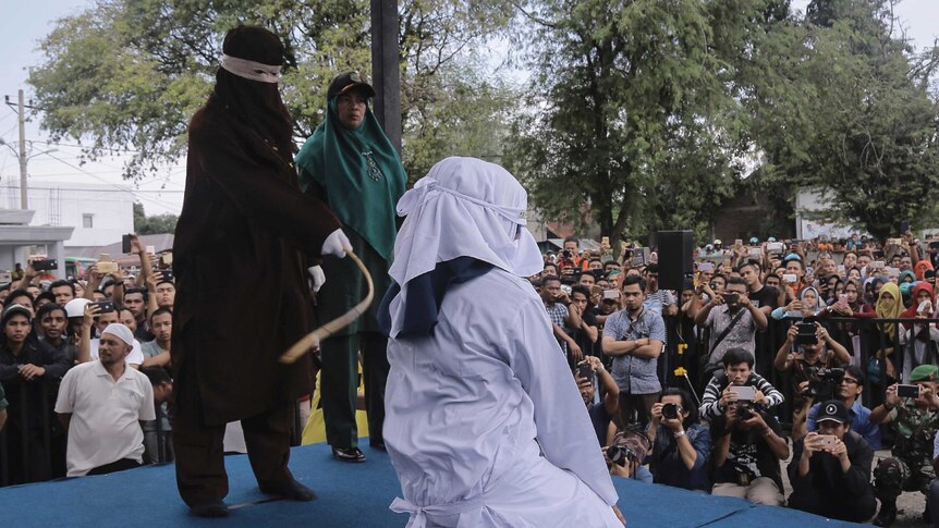 Wide shot of a person in black robes whipping a person in white robes.