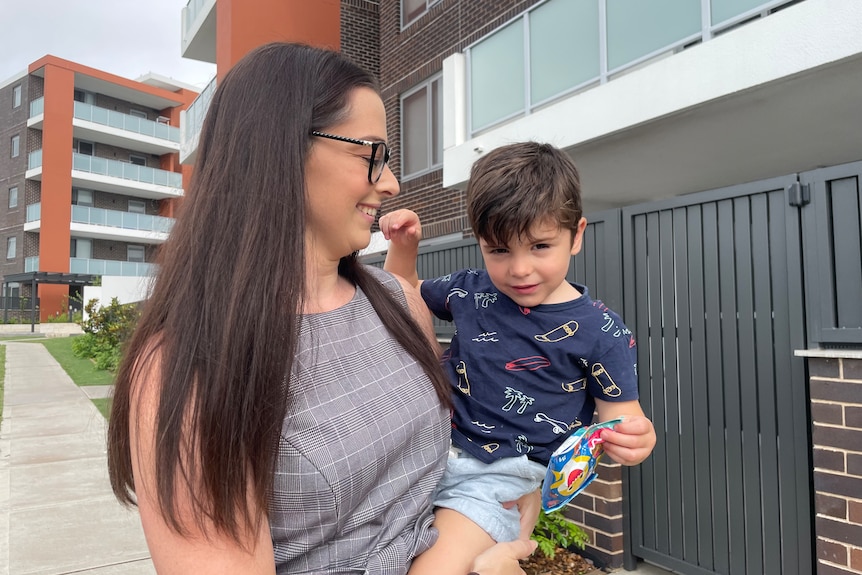 Mother holding toddler standing outside an apartment block.