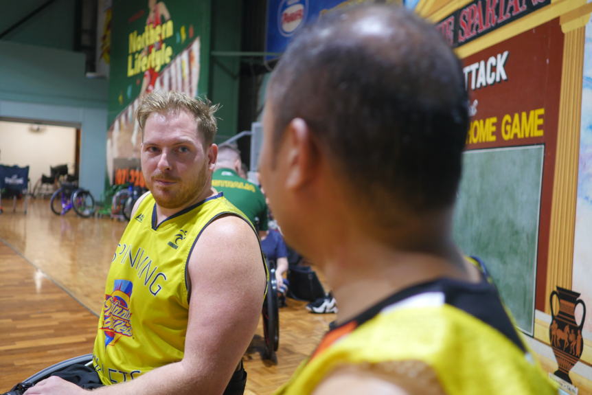 Two men using wheelchairs, talk together on the side of a basketball court.