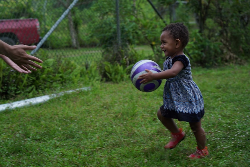 A toddler runs through the grass clutching a ball