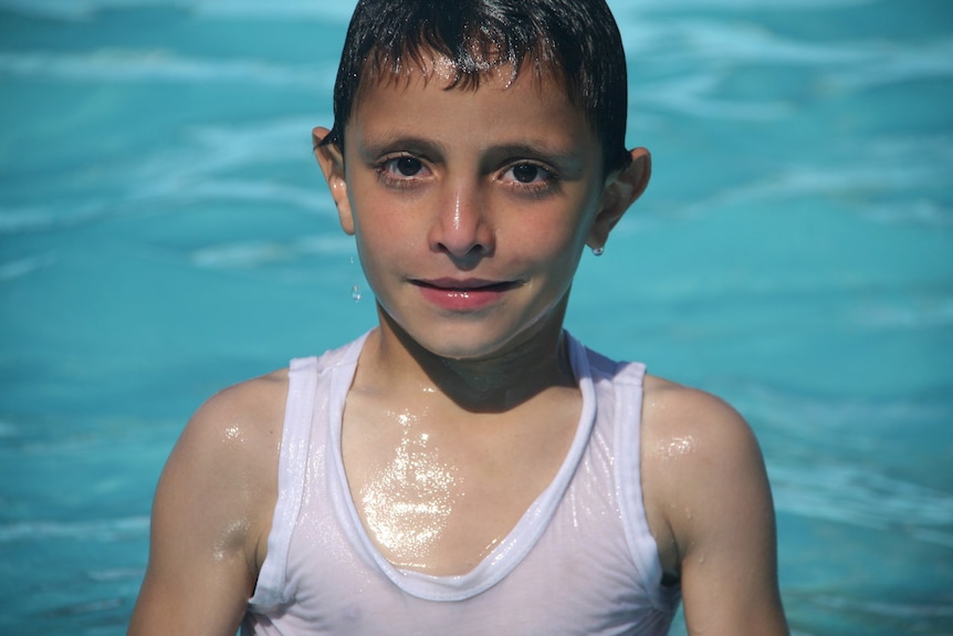 A close up of a young boy who is wet and wearing a singlet, standing in the ocean and looking directly at the camera.
