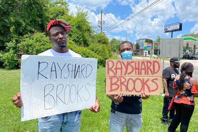 Two black men hold signs with the name Rayshard Brooks, standing on a grassy area on sunny day.