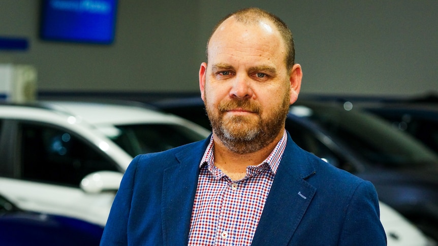 A man in a button-up shirt and blazer stands in front of a row of electric vehicles.