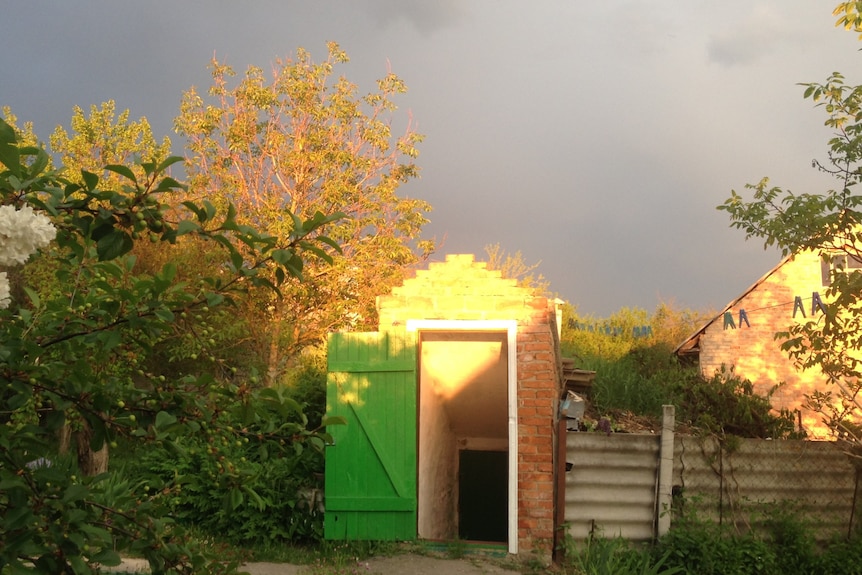 A bomb shelter with a green door in the backyard of a small village surrounded by trees with grey skies in the background