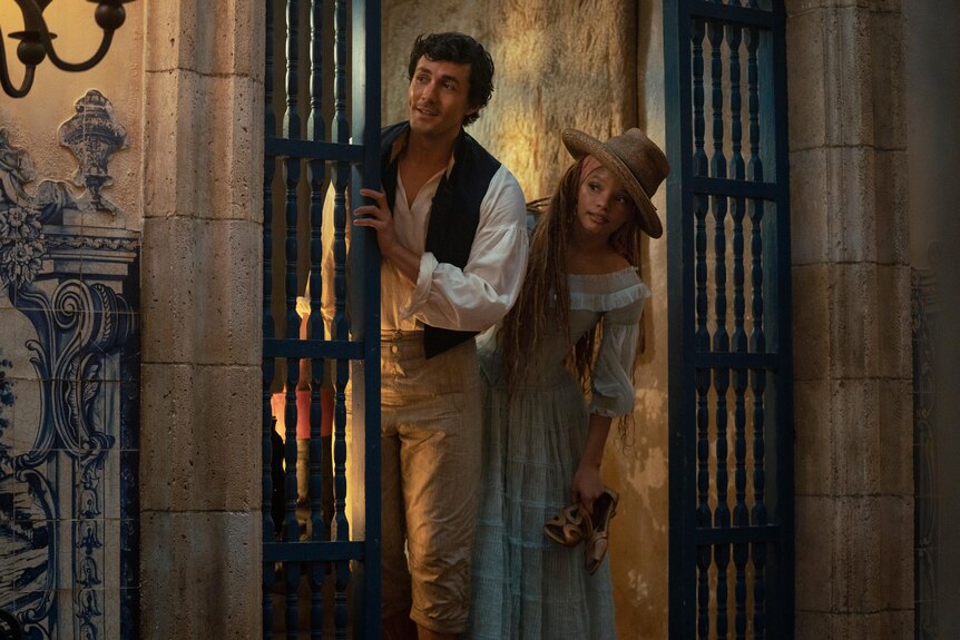 A man and a woman in old-fashioned clothing peek around the corner of a gate, ina