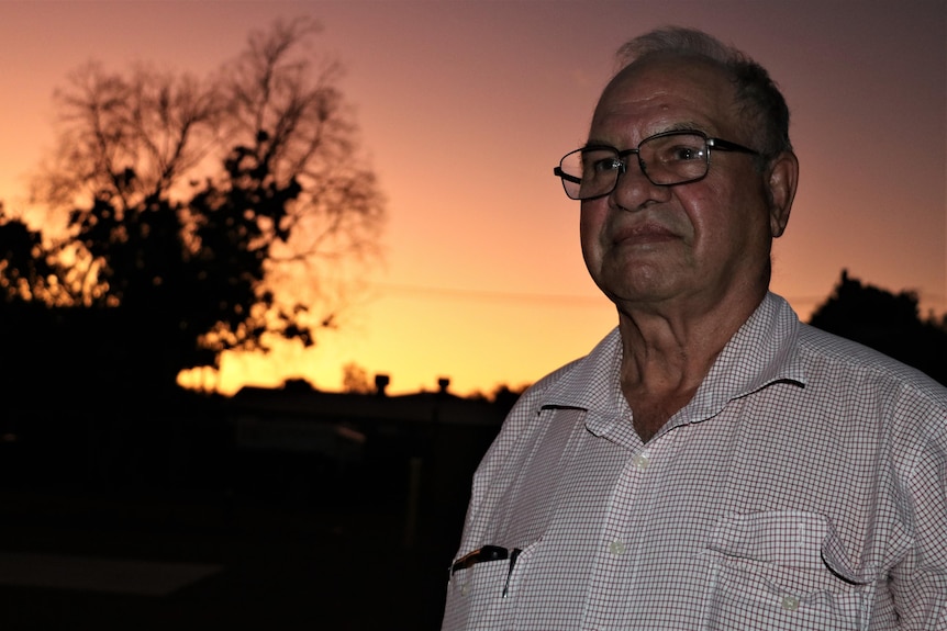 An older man wearing a white checked shirt stands looking off into the distance with an orange and yellow sunset behind them
