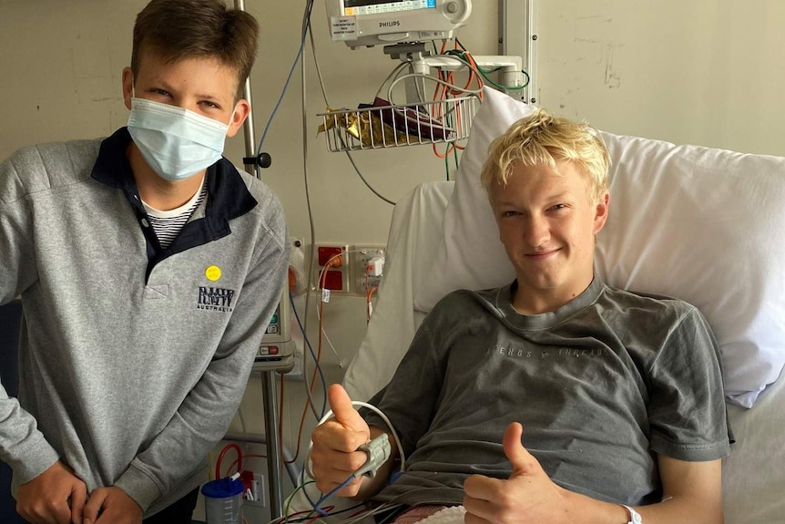 Boy in mask standing on left of hospital bed, blond boy in bed smiling with thumbs up