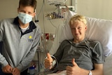 Boy in mask standing on left of hospital bed, blond boy in bed smiling with thumbs up