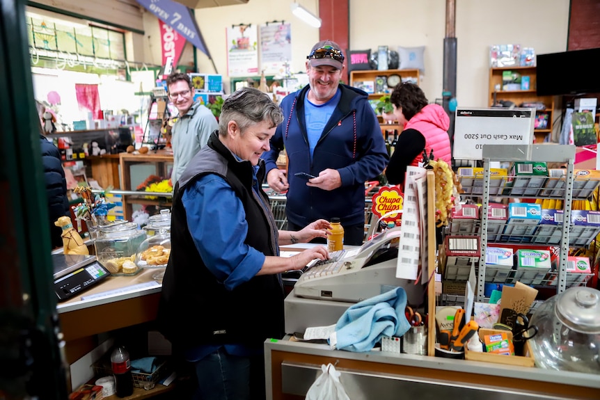 Woman in blue shirt and vest stands at shop cash register with three smiling customers waiting inside general store.