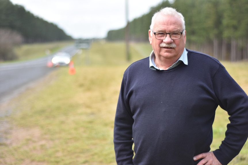 A white-haired man in a navy jumper stands by the side of the highway with grass and a forest behind him