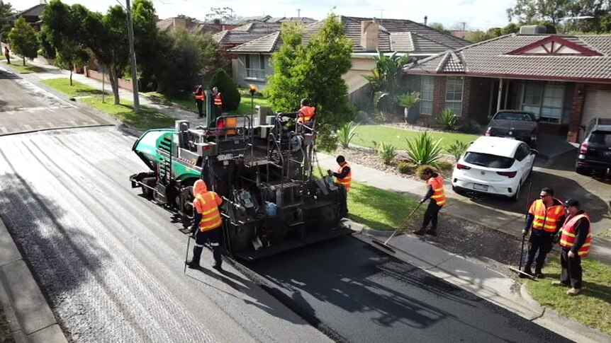 Workers dressed in hi-vis vests work on a suburban road as a large vehicle lays asphalt.