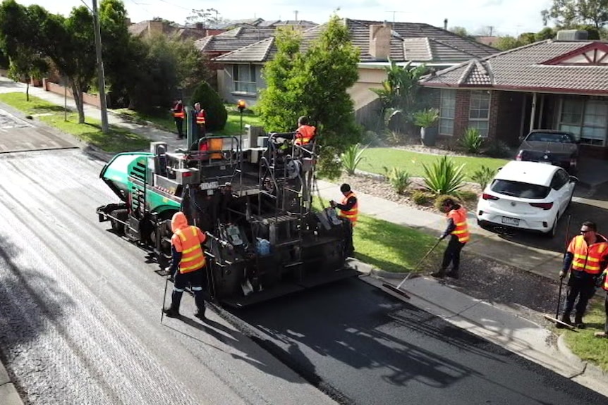 Workers dressed in hi-vis vests work on a suburban road as a large vehicle lays asphalt.