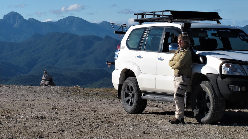 Woman leans against white four wheel drive, mountains in background