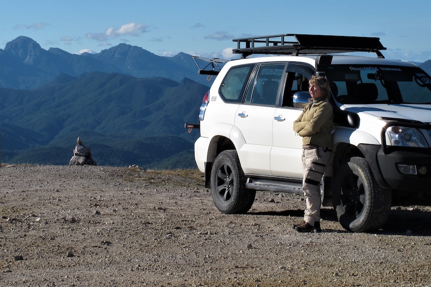 Woman leans against white four wheel drive, mountains in background