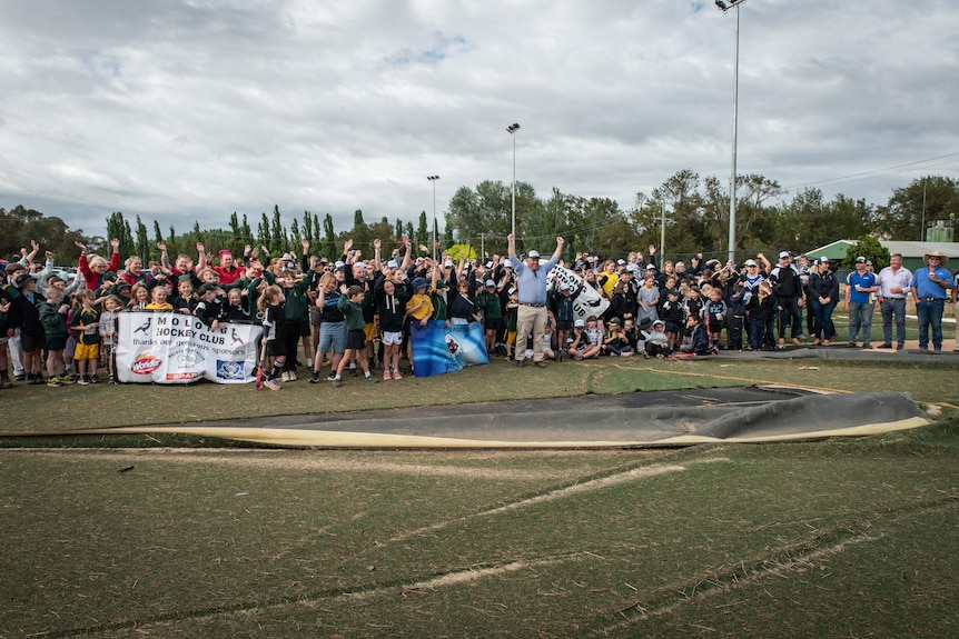 A big group of children and adults stand together cheering on a flood ruined hockey oval.