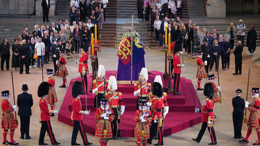 People in queues watch royal guards marching around and standing by the Queen's coffin.