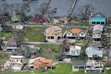 Houses by a lake are seen with pieces of roof and missing and debris is scattered on the ground.