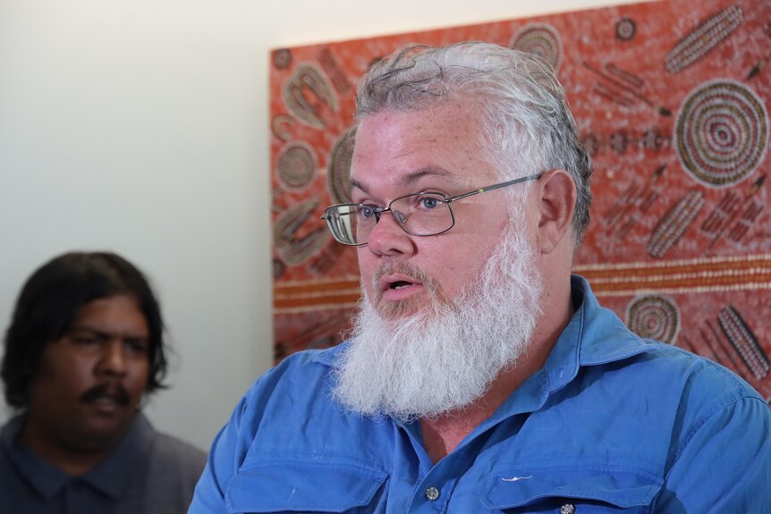 Man with glasses and beard stands in front of Aboriginal art and speaks