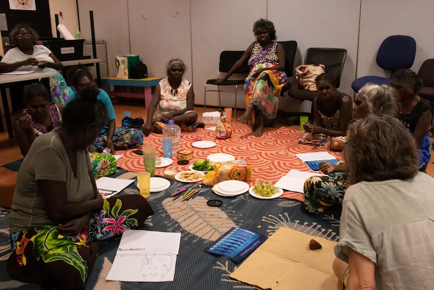 Women sit in a circle inside. In the middle is paper and snacks.